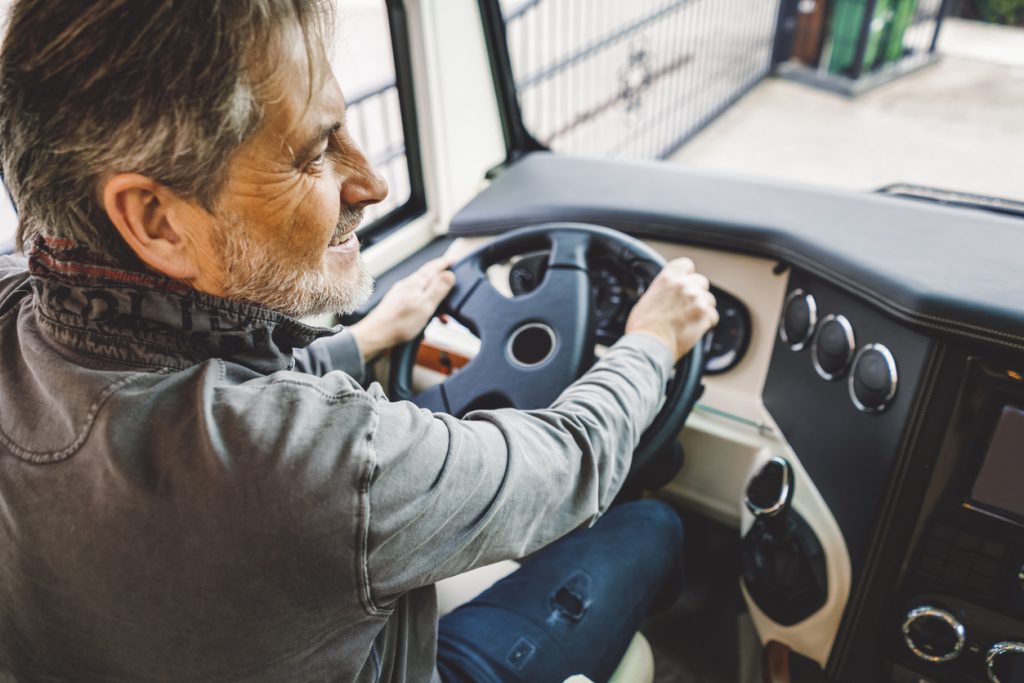 Driving a motorhome - Mature man driving a camper, happy man having grey hair and a beard, wearing grey hoodie and dark blue trousers, holding a steering wheel, feeling happy, going on a trip, it's vacation time, road trips are the best.