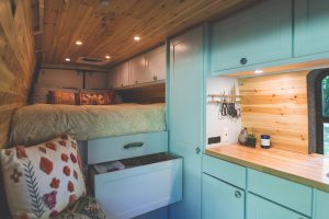 right RV furniture - Interior of a van that a young couple live in. The shot is focused on the kitchen counter, pull-out drawers, and bed. Ther is wood paneling on the sides and roof.