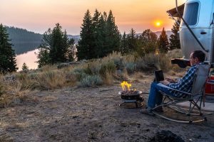 A man on his laptop outside of his travel trailer with a campfire and a scenic view. 
