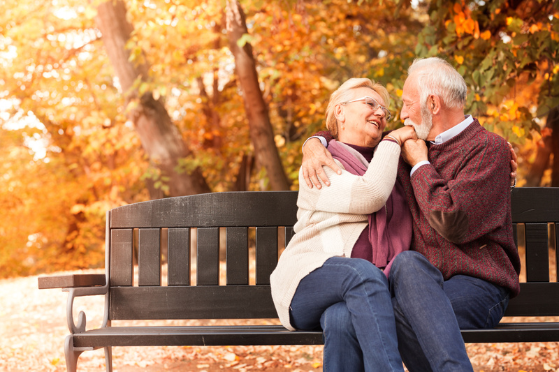 A woman smiles as her husband kisses her hand on a park bench among orange autumn leaves.