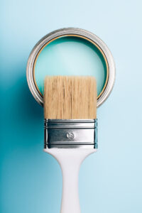 Brush with white handle on open can of turquoise paint on blue pastel background